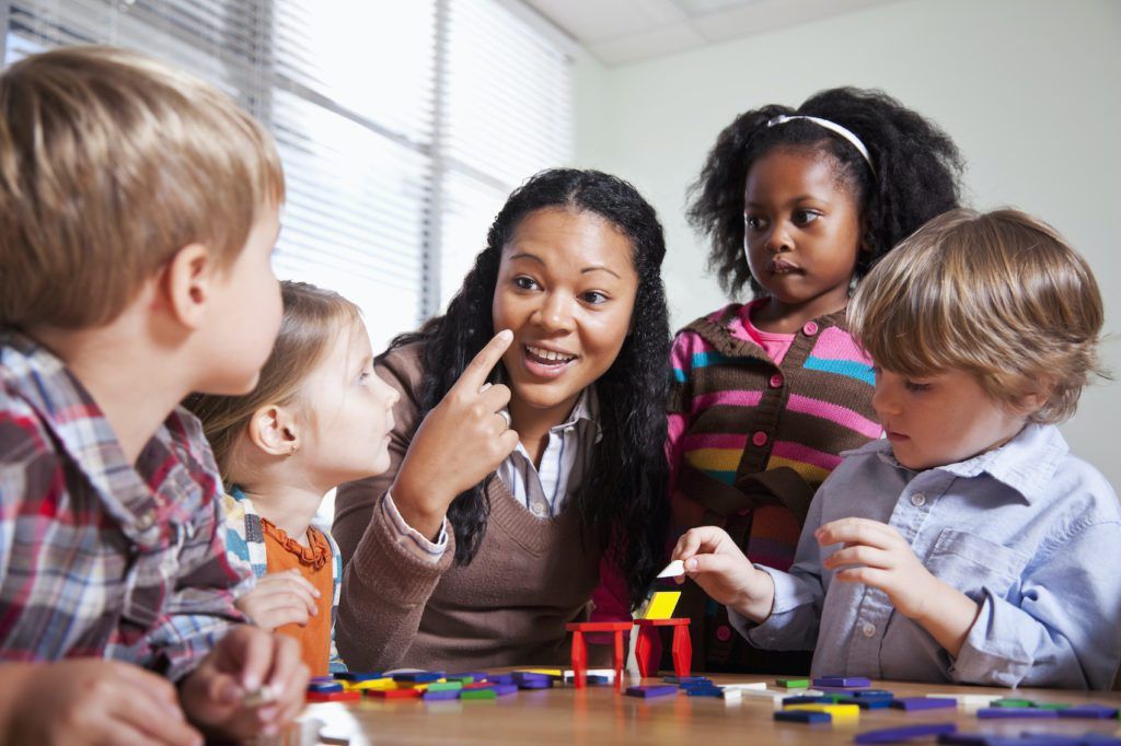 A teacher listening warmly to a small group of children gathered around a classroom table.